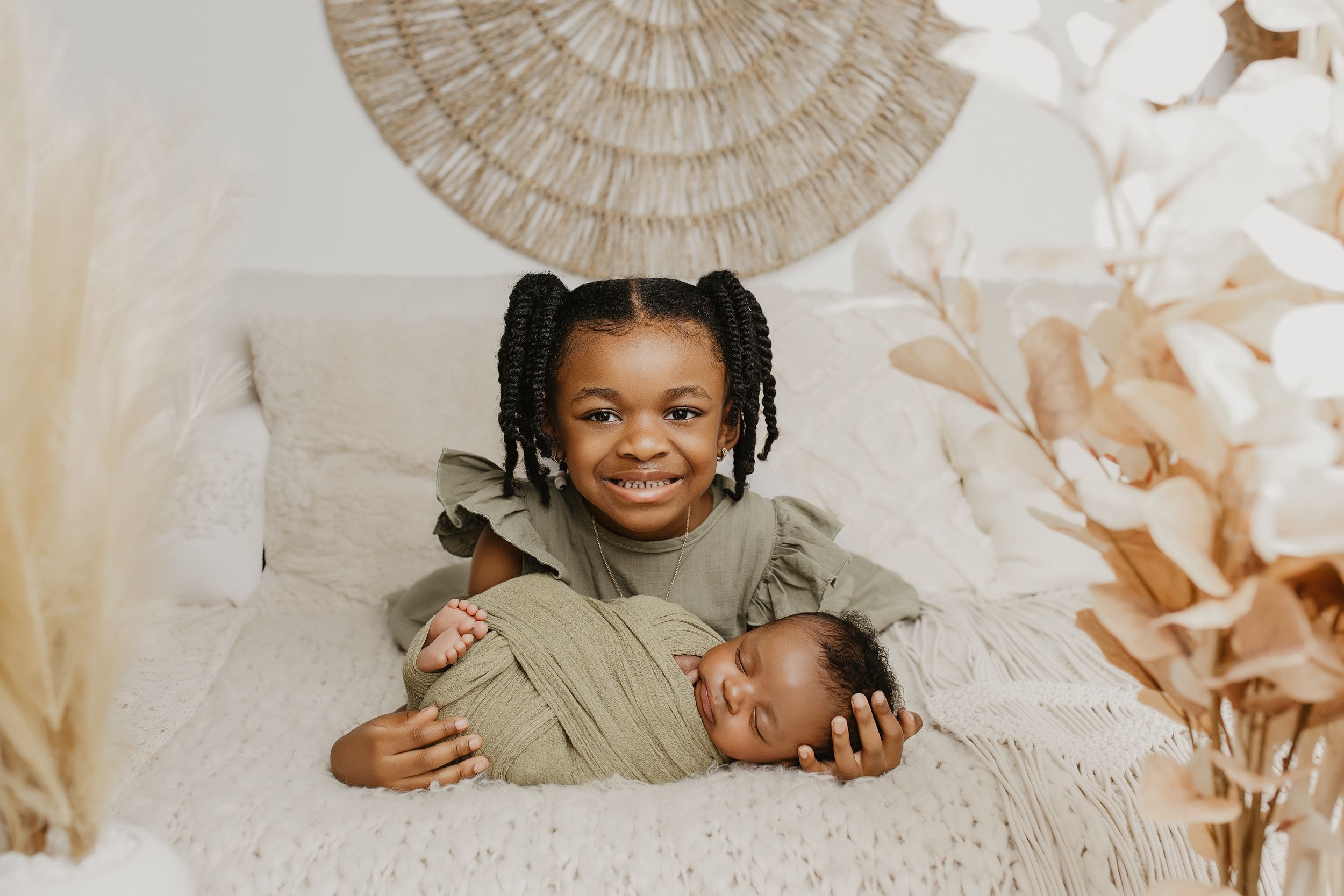 Smiling child with braids poses with sleeping newborn baby on white bed with boho decor.