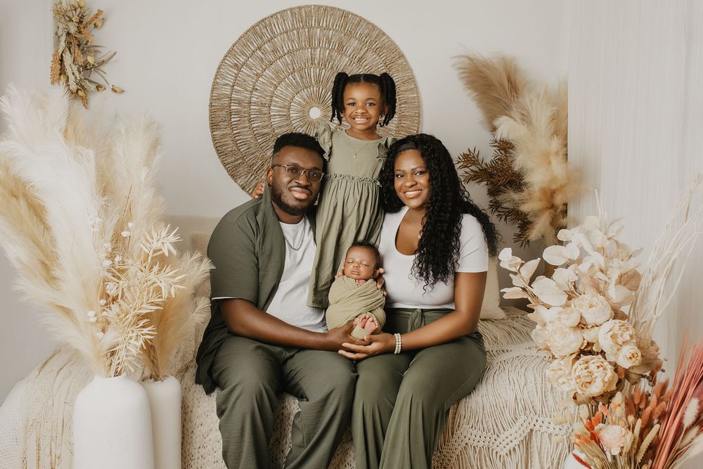 A joyful Black family of four poses together in a boho-styled photo studio with pampas grass and floral decor.