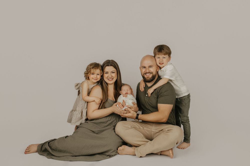 Family of five poses for a studio portrait, parents sitting on floor with newborn baby and two young children in neutral tones.