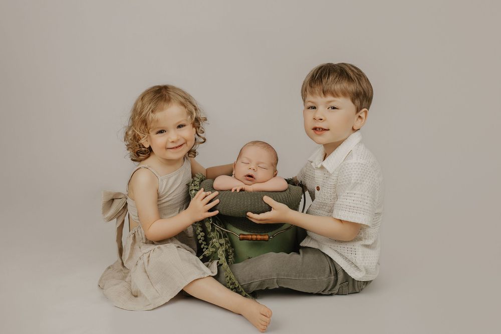 Two young siblings sit together holding a sleeping newborn baby in a green bowl during a studio photo session.
