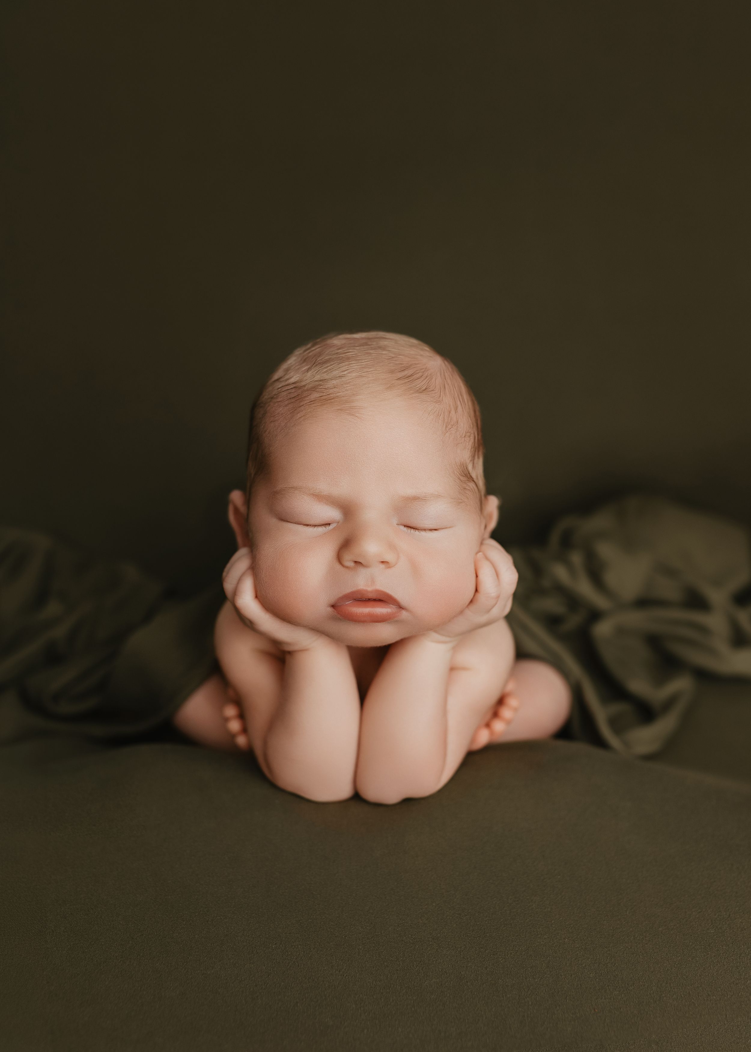 Sleeping newborn baby posed with hands under chin on olive green backdrop in professional newborn photography session.