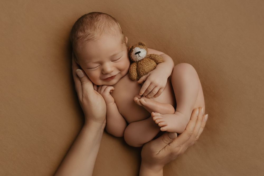 Smiling newborn baby curled up holding a tiny teddy bear, cradled in parents hands on a warm brown background.