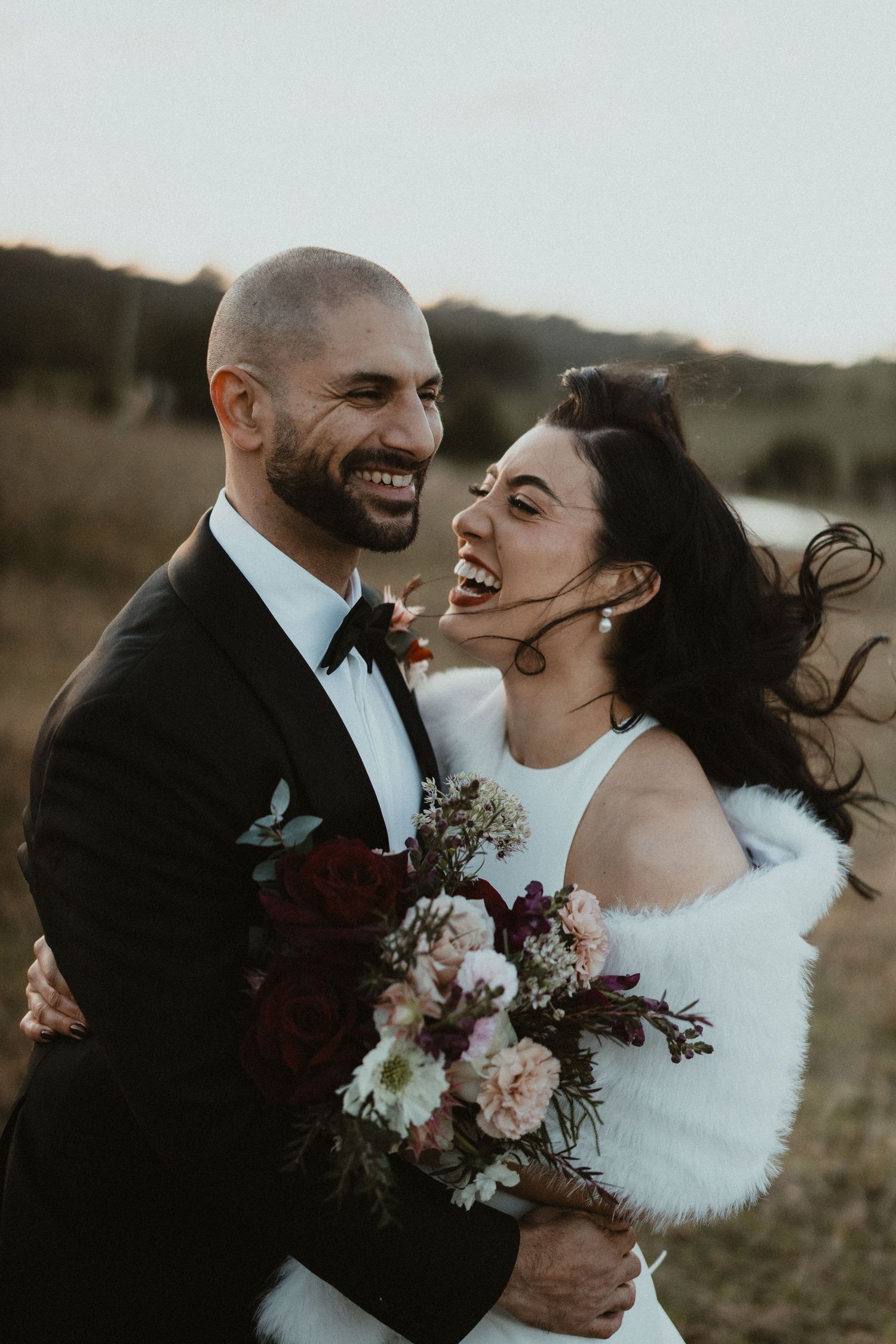 Newlywed couple sharing a candid laughing moment with bridal bouquet at sunset.