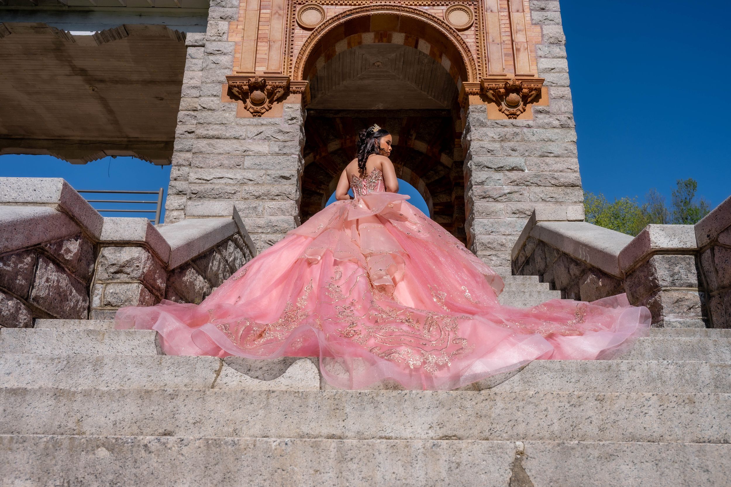 Young woman in a stunning pink ball gown quinceañera dress poses on stone steps of an elegant building.