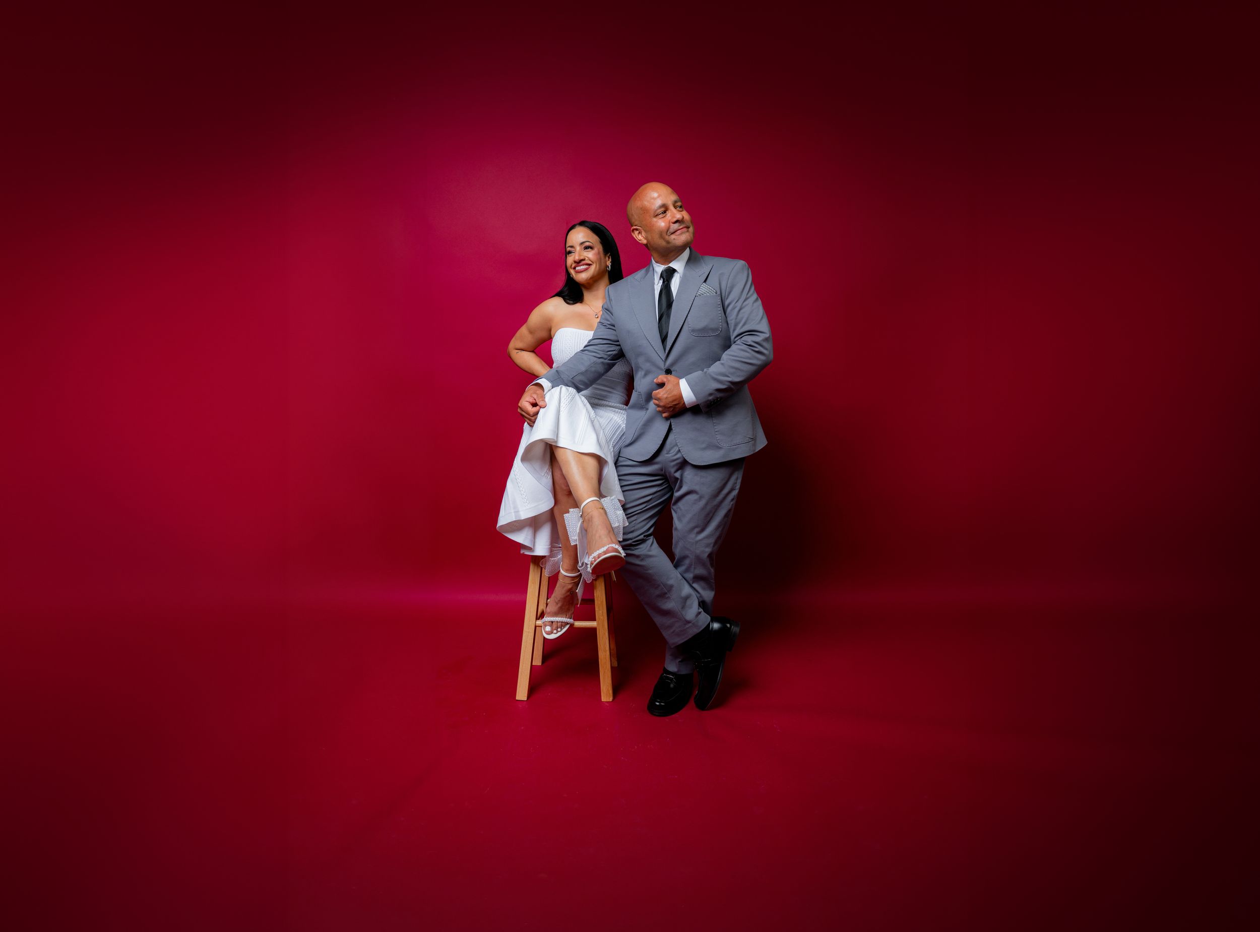 Wedding couple poses creatively in studio with deep red background wearing grey suit and white dress.