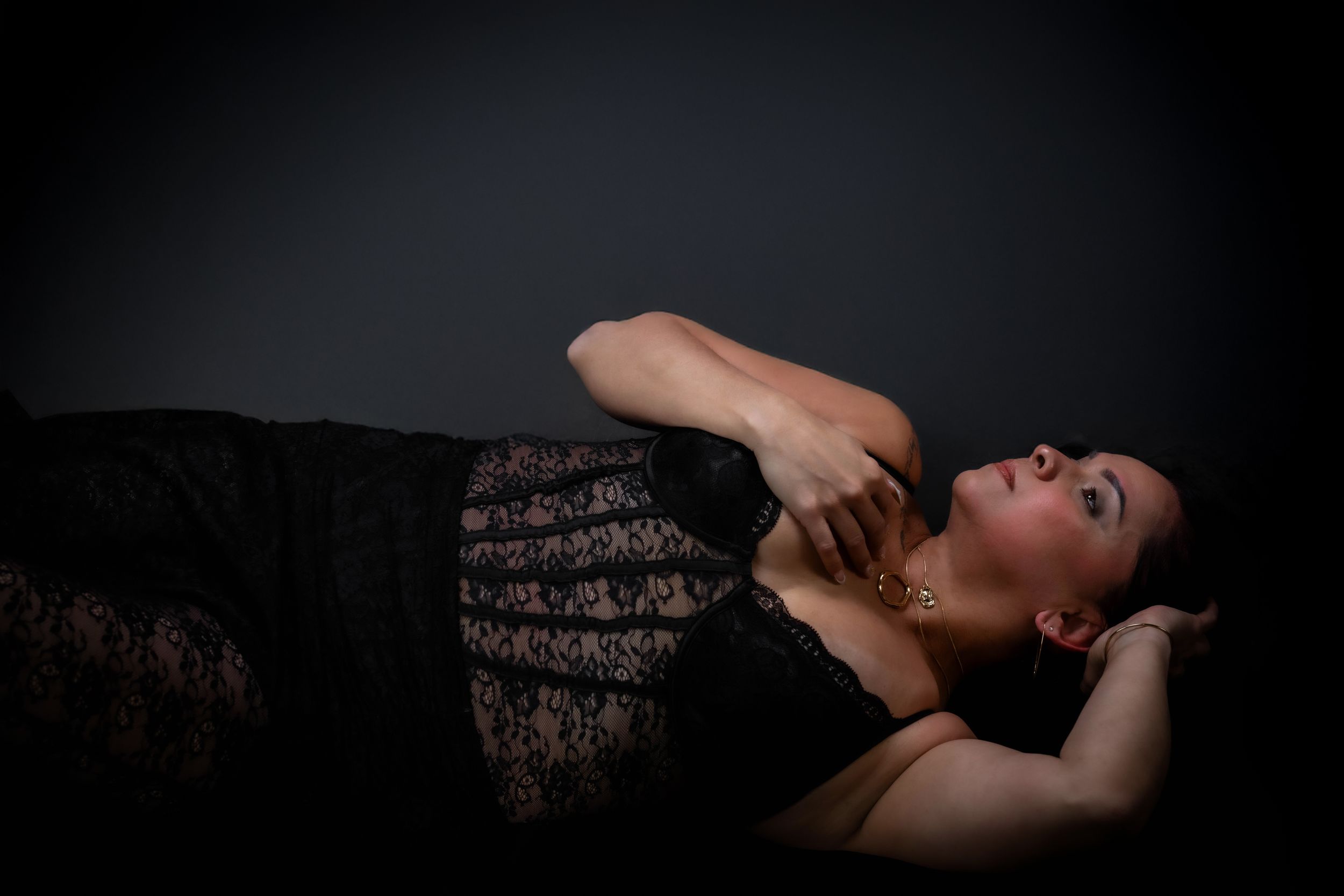 Woman in black lace dress lying down elegantly against dark background in artistic boudoir photography.
