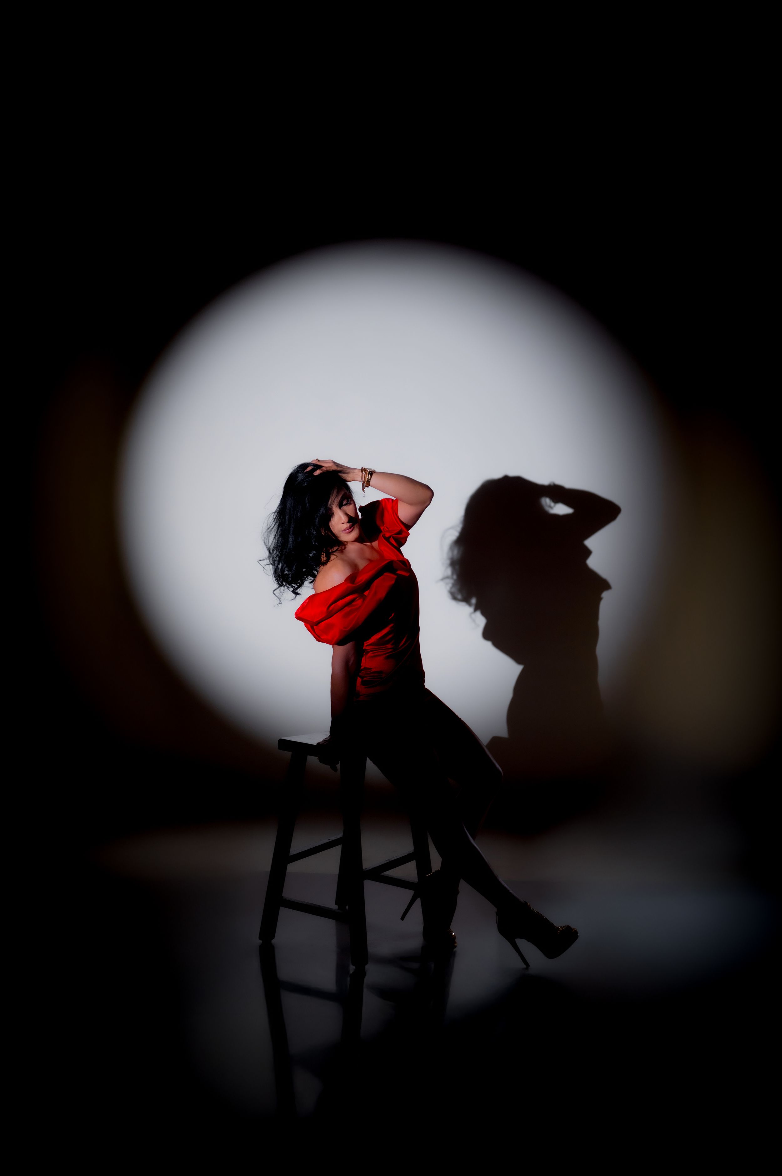 Woman in red top seated on stool, lit by spotlight with dramatic shadow on white background.