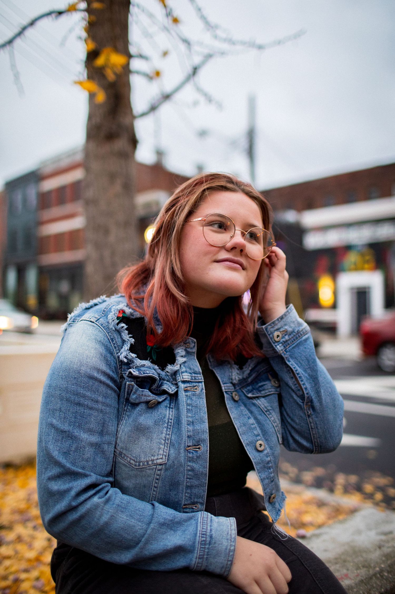 A young woman with pink hair and glasses sits outdoors in a denim jacket among autumn leaves.