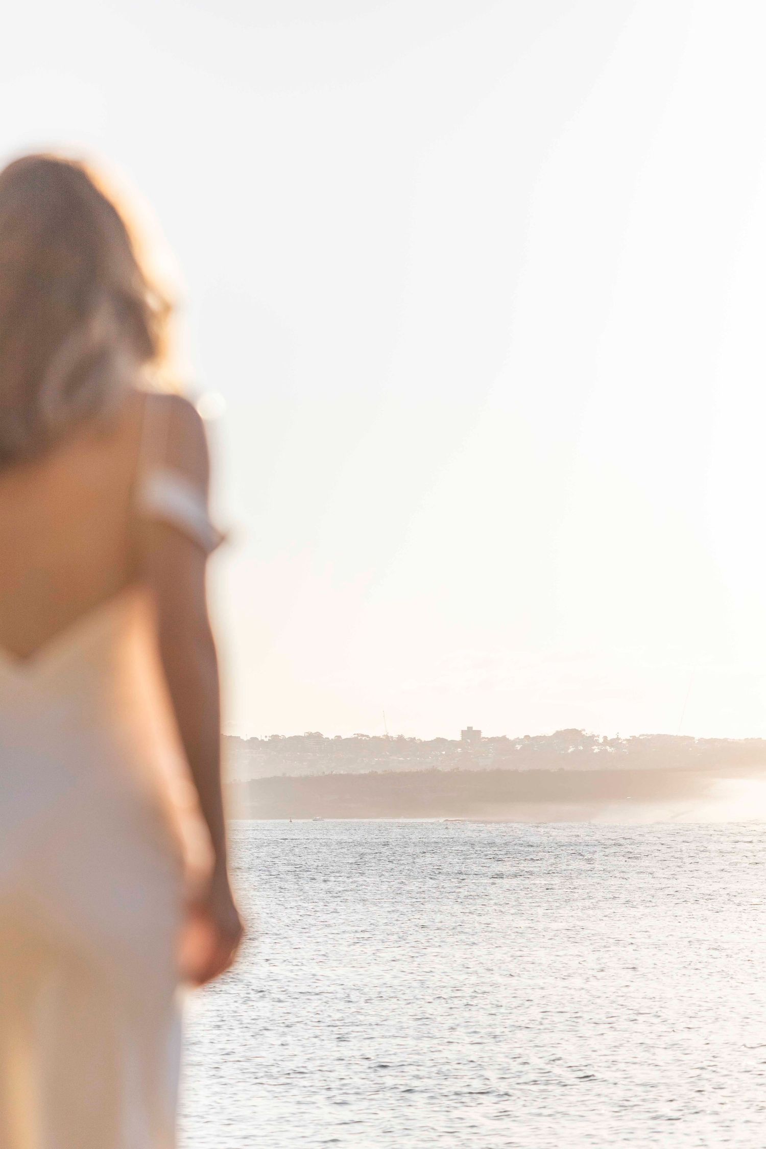 Blurred silhouette of someone in white dress looking towards water at sunset.