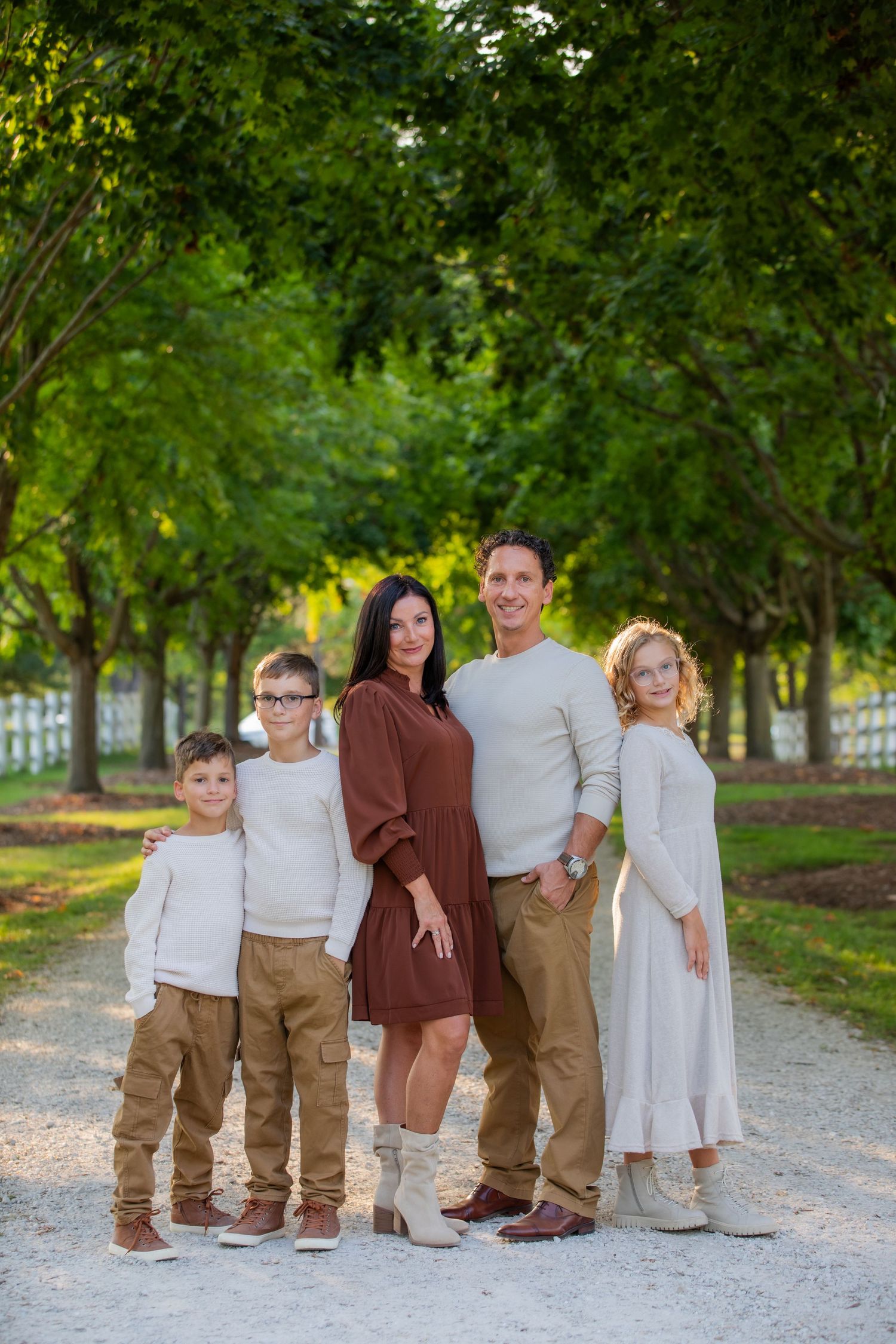 A family stands together on a gravel path surrounded by green trees in coordinated brown and white outfits.
