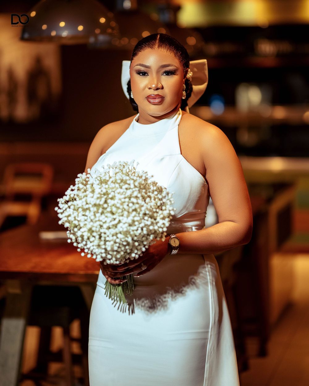A person in a white sleeveless dress holds a delicate bouquet of baby's breath flowers against a warm-lit background.