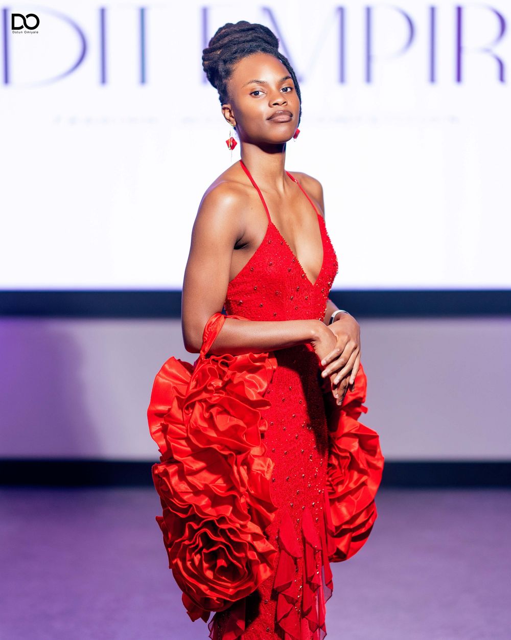 Fashion model in red evening gown with elaborate ruffled skirt poses on purple-lit runway.