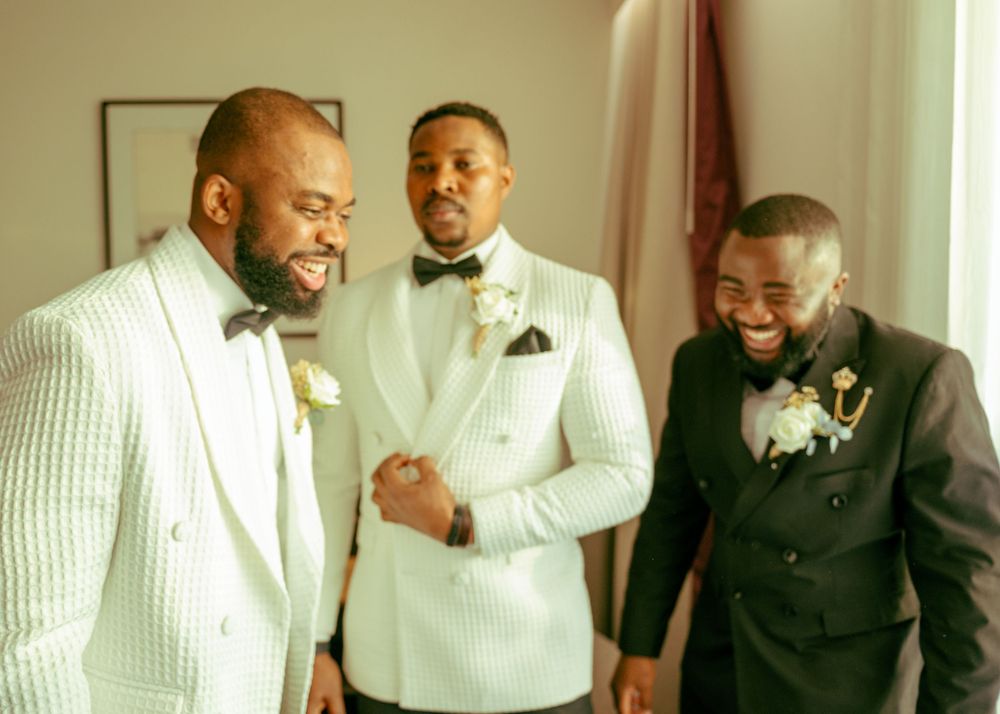 A group of men in formal white tuxedos and black bowties share a laugh together during a wedding celebration.