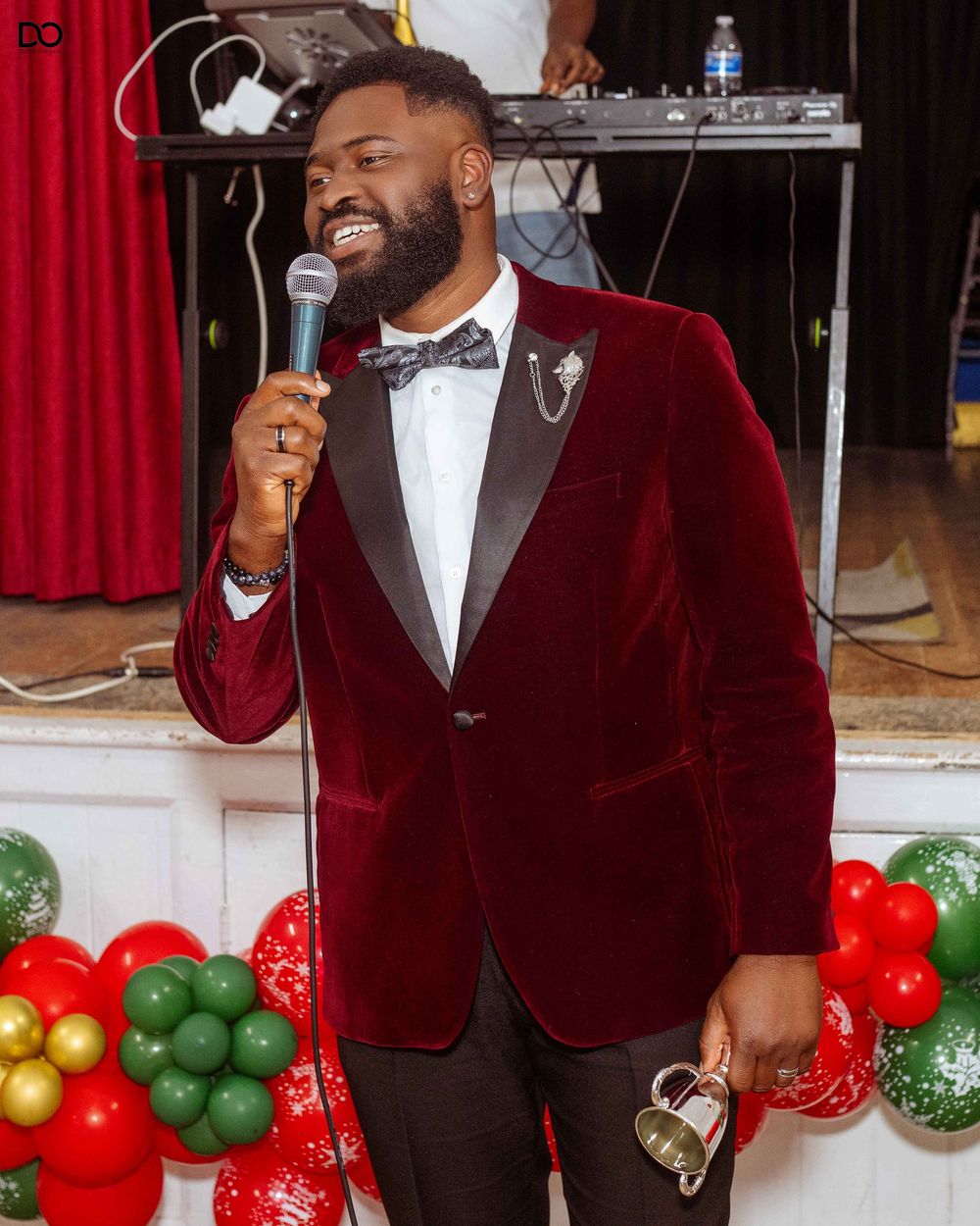 A performer in a burgundy velvet blazer and black bow tie sings passionately on stage with festive holiday balloons.