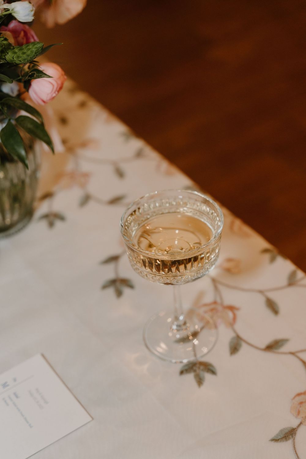 glass of champagne. Champagne in vintage glass on table. Tablecloth with pink flowers. Wedding tablescape. Vintage wedding table with drinks