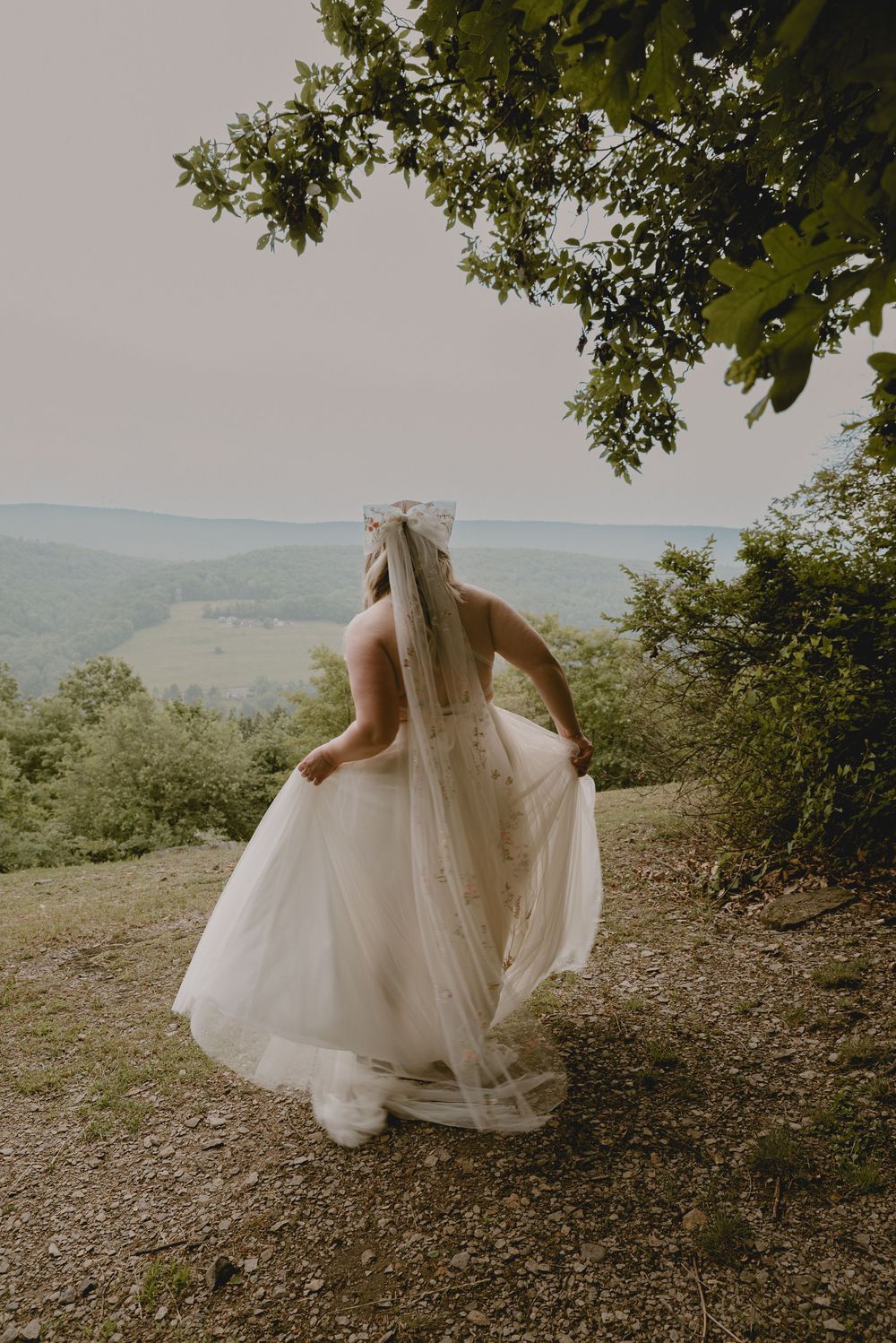 bride standing on mountain with a view in the background holding her dress. Bow veil flow bridal gown. Bride looking out to view. Bride bow