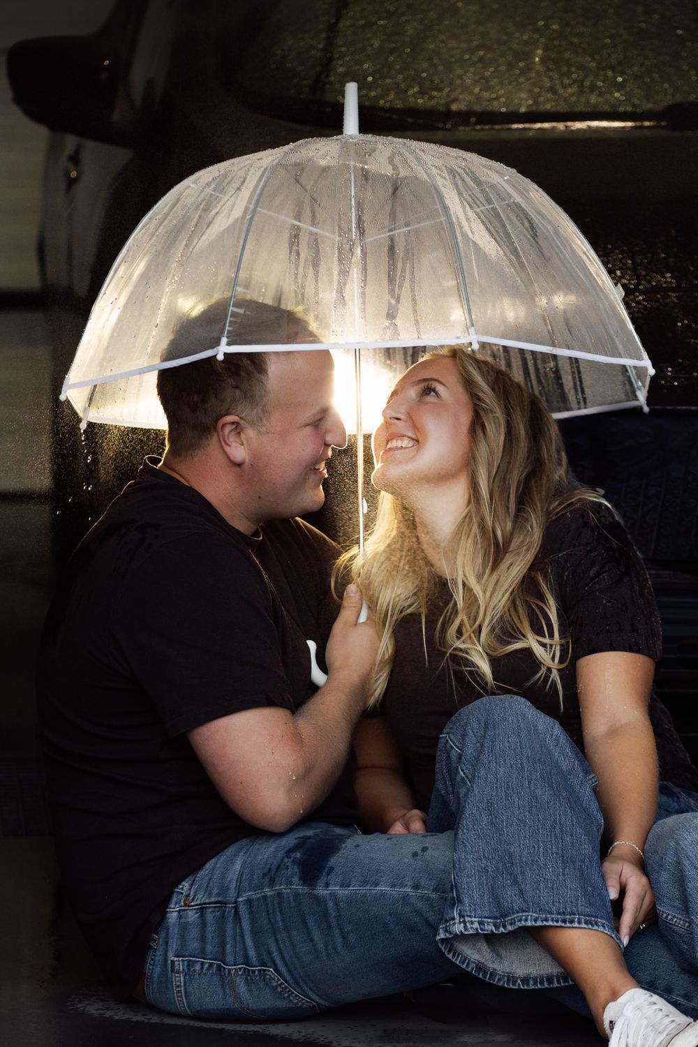 Couple sitting together under a clear umbrella in the rain, smiling at each other while wearing matching black shirts and jeans.