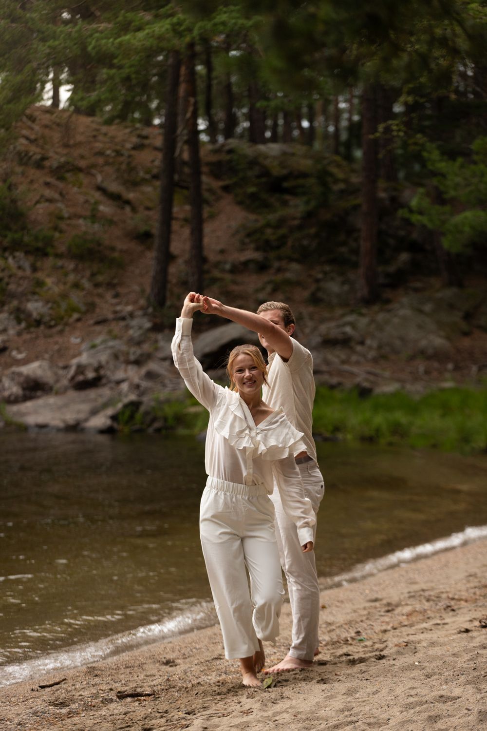 Couple in white clothing dances together on lakeside beach.