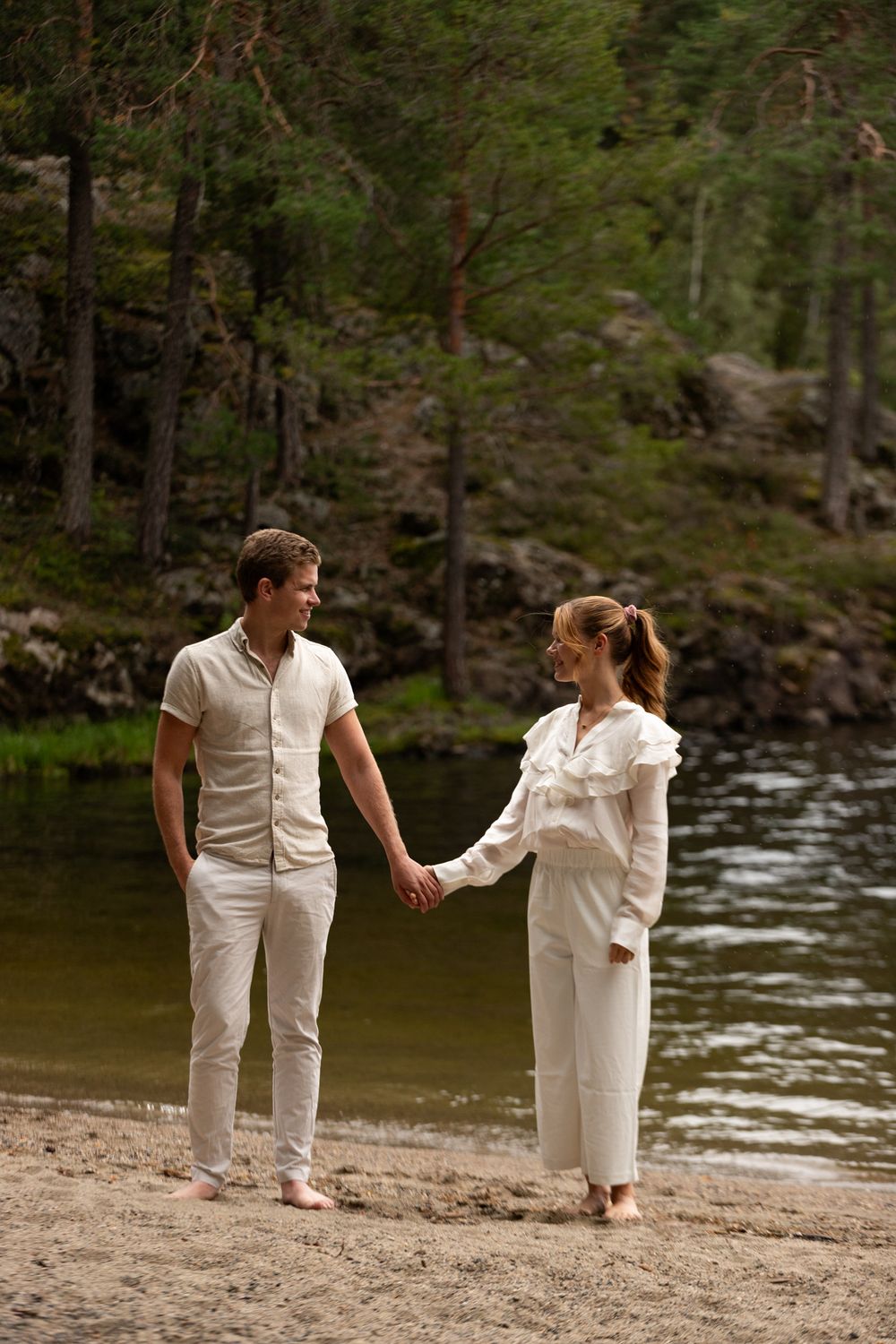 Couple in matching white outfits shares intimate moment on beach near lake.