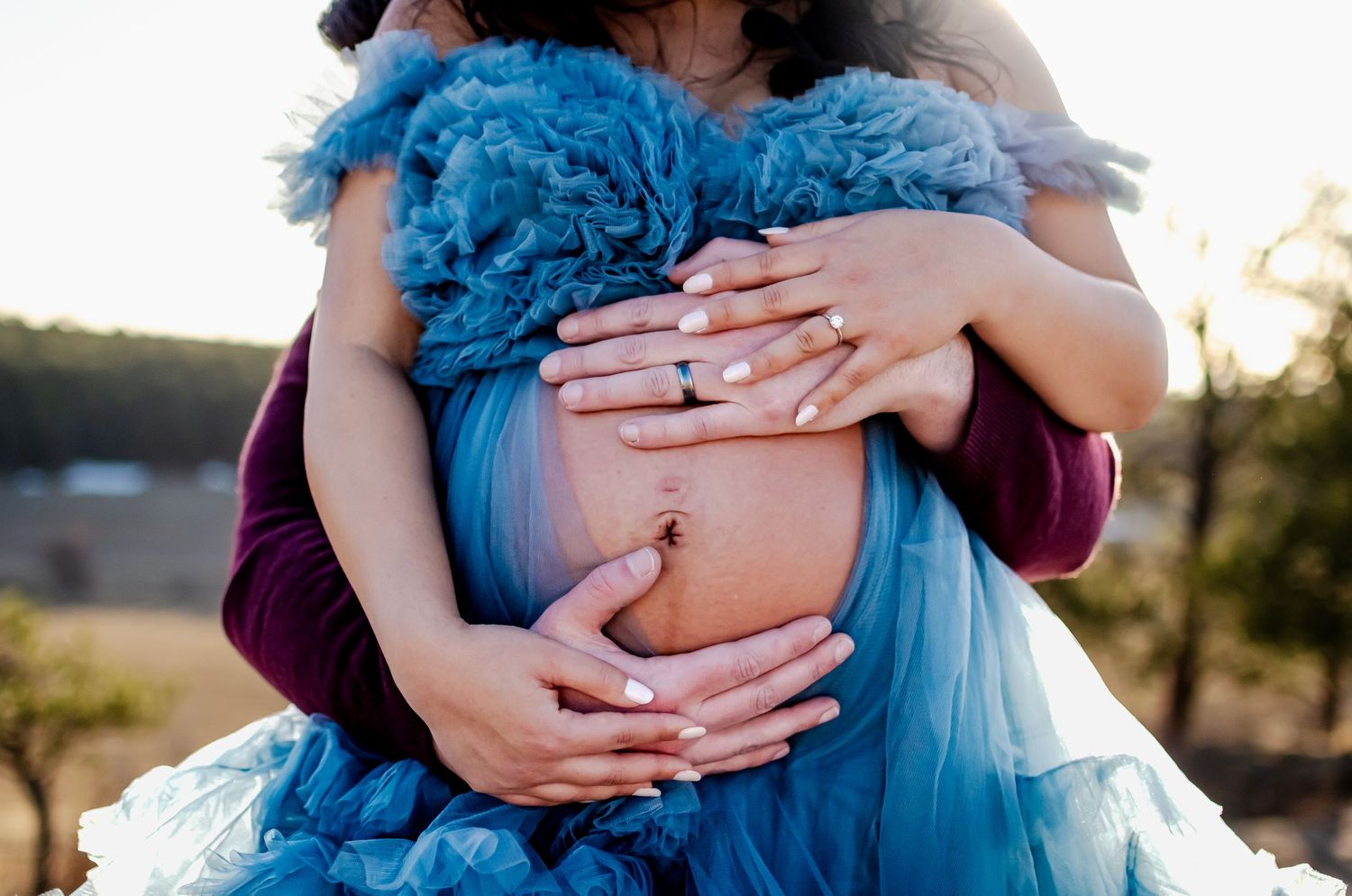 A close up of hands cradling a baby bump through blue tulle fabric at sunset.