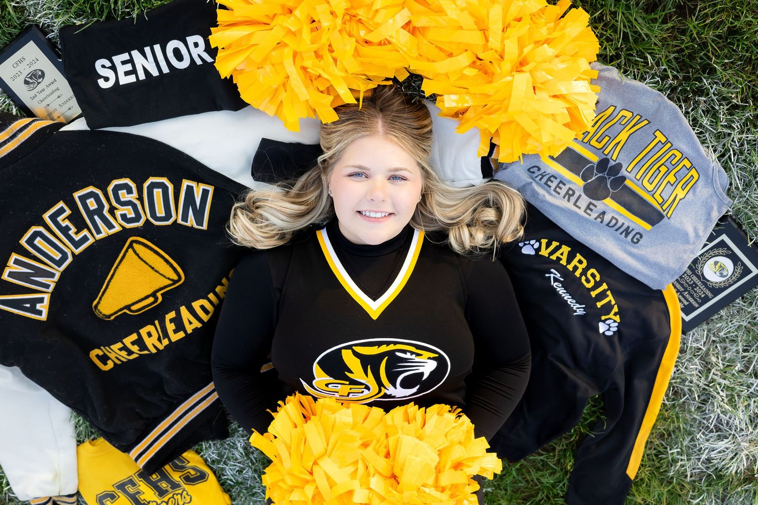 A cheerleader in black and gold uniform lies on grass surrounded by pom poms and varsity jackets.