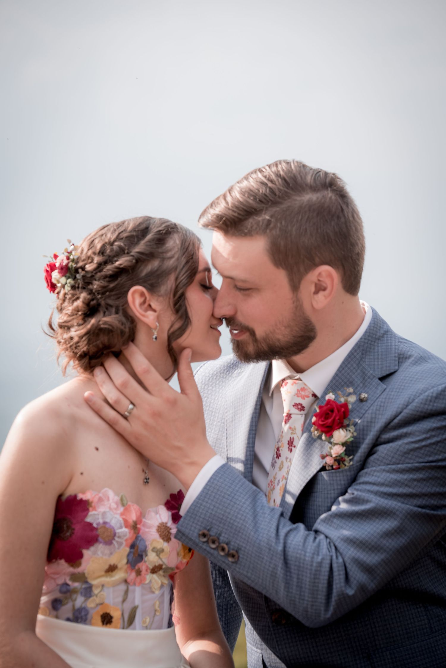 Romantic wedding couple share an intimate moment in formal attire with red floral accents against soft gray background.