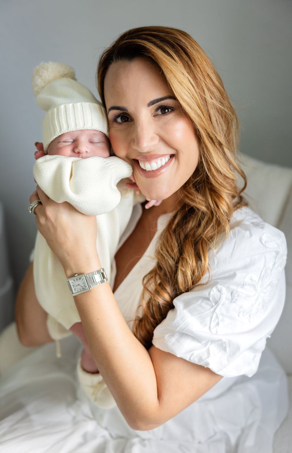 Smiling mother in white dress holds sleeping newborn in cream knit hat close to her cheek in a bright, airy room.
