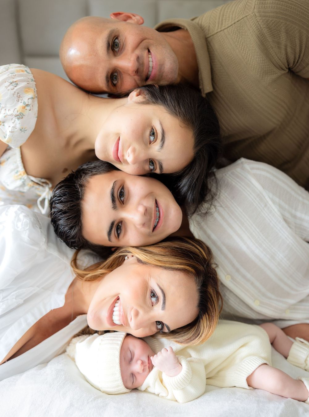 Family of five posing together on a bed, smiling warmly, with a newborn baby in front.