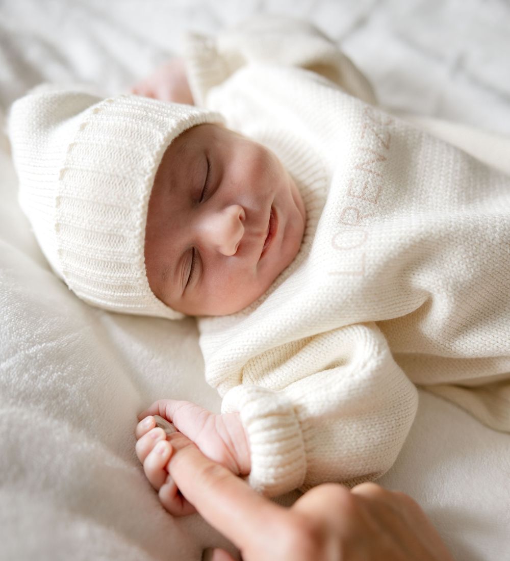 Sleeping newborn baby in cream knit outfit and hat, cradled gently in adult hands on soft white blanket.