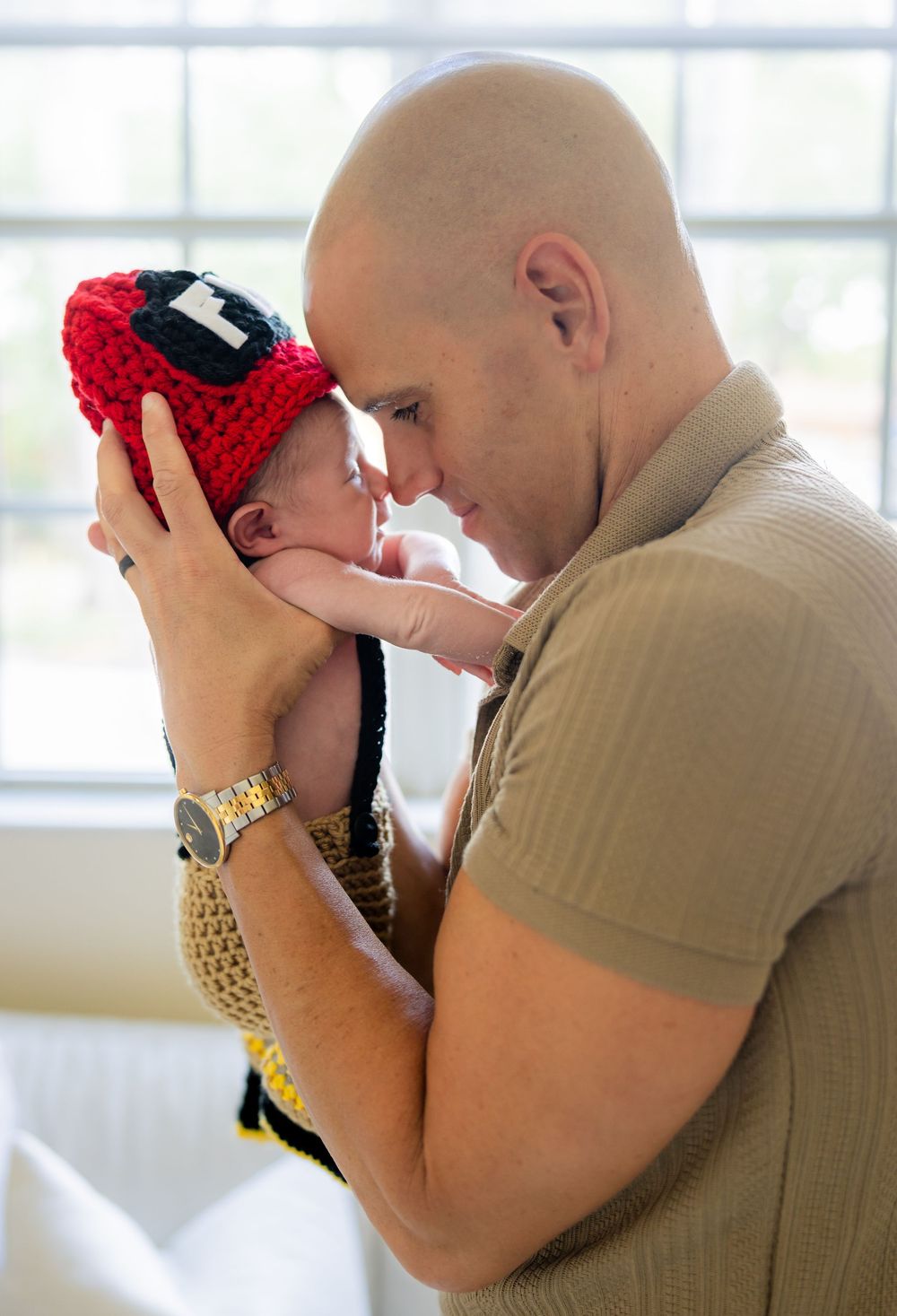 Bald man tenderly holds newborn baby wearing red crocheted firefighter hat, touching foreheads in loving embrace.