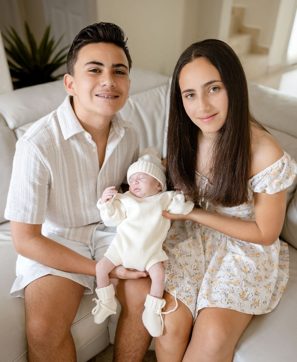 Two teenagers smiling and holding a newborn baby dressed in white, sitting together on a white couch.