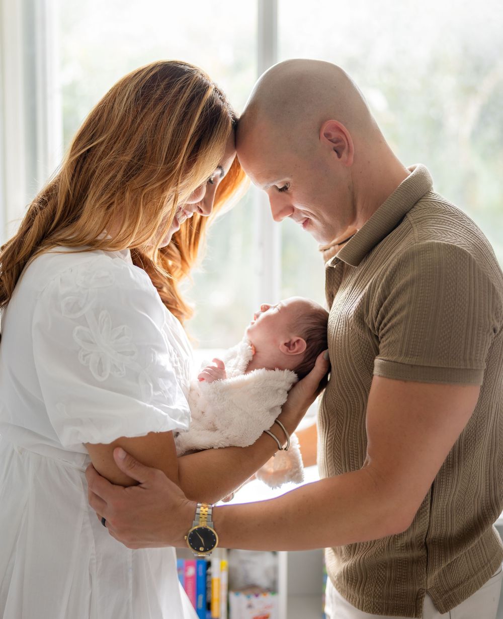 A couple tenderly gazes at their newborn baby, the mother in white dress and father in tan polo, sharing a precious family moment.