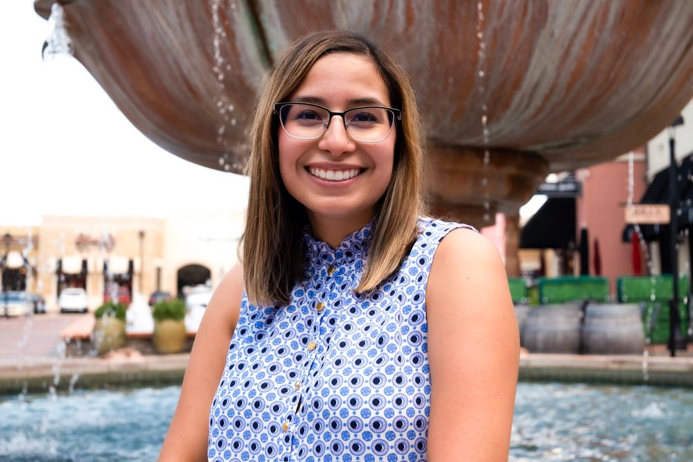 Someone in glasses and a sleeveless polka dot blouse smiles while standing in front of an outdoor fountain.