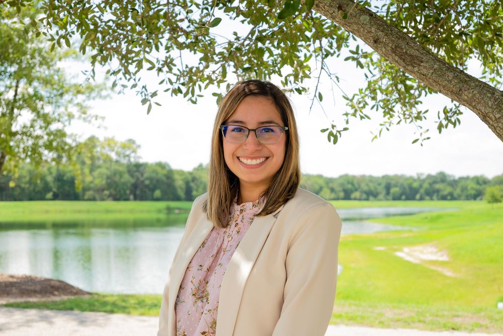 Woman in white blazer and sunglasses poses in a sunny outdoor setting with green grass and trees in background.