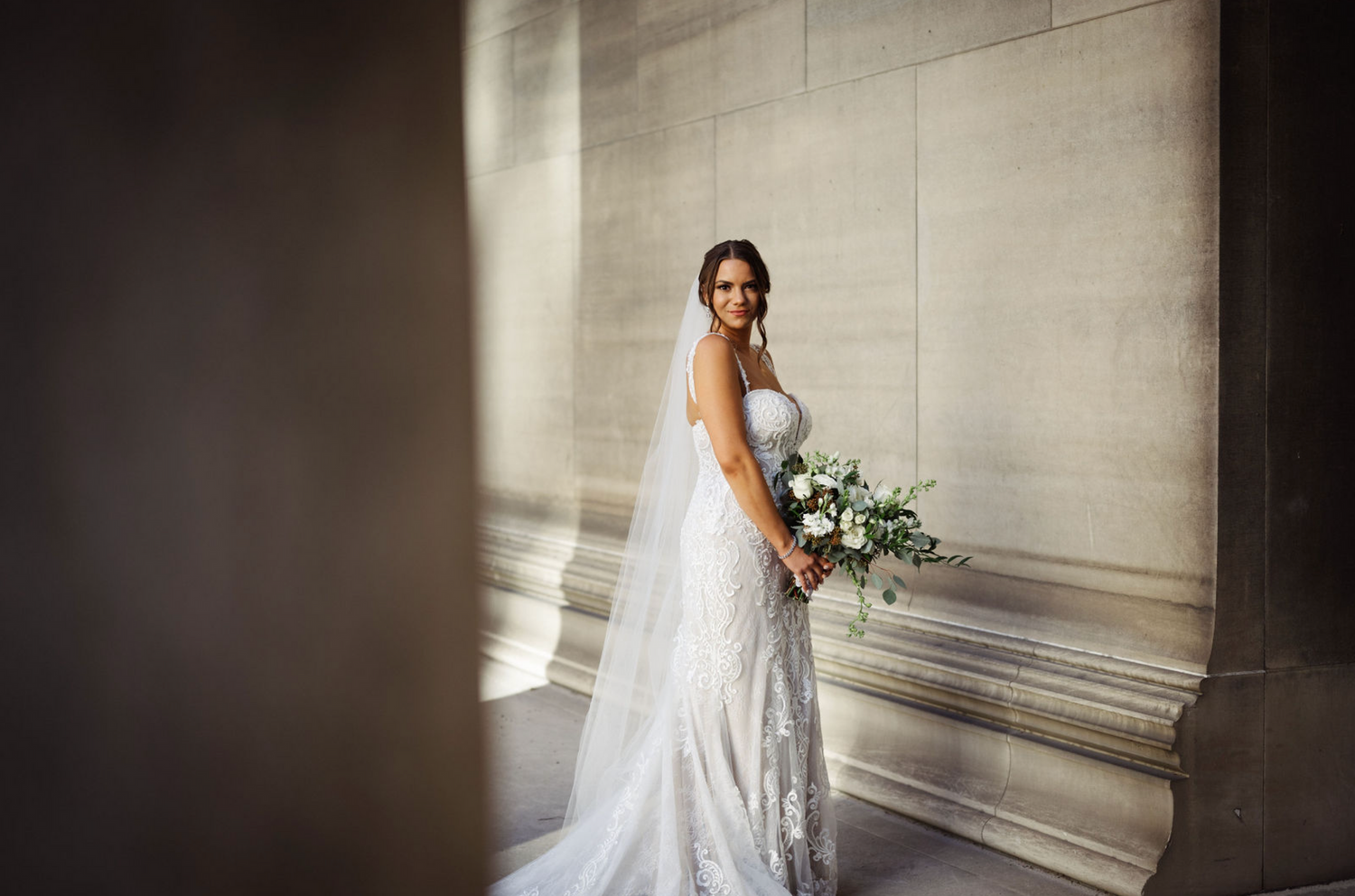 Elegant bride in lace gown holding white bouquet poses between stone columns in a stunning bridal portrait.
