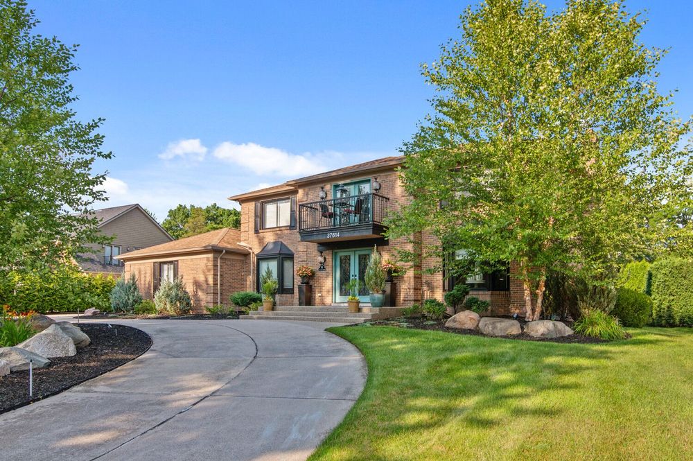 Two-story brick home with curved driveway, lush landscaping, mature trees, and a welcoming front entrance.