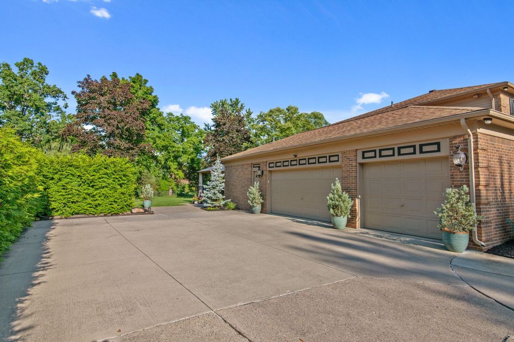 Spacious concrete driveway leading to a two-car attached garage surrounded by mature trees and hedges.