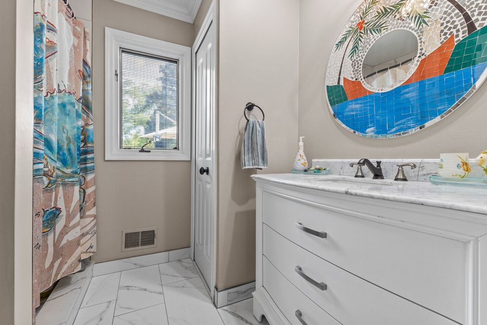 Bright modern bathroom with white vanity, marble-look tile floors, colorful round mirror, and walk-in shower.