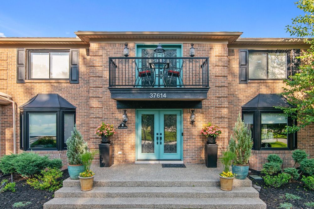 Brick home exterior with teal double entry doors, balcony, black awnings, and manicured landscaping at entrance.