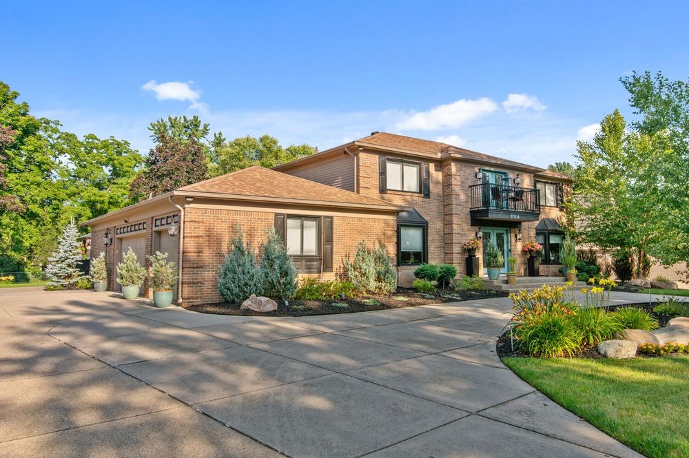 Two-story brick and wood home with circular driveway, attached garage, balcony, and mature trees in background.