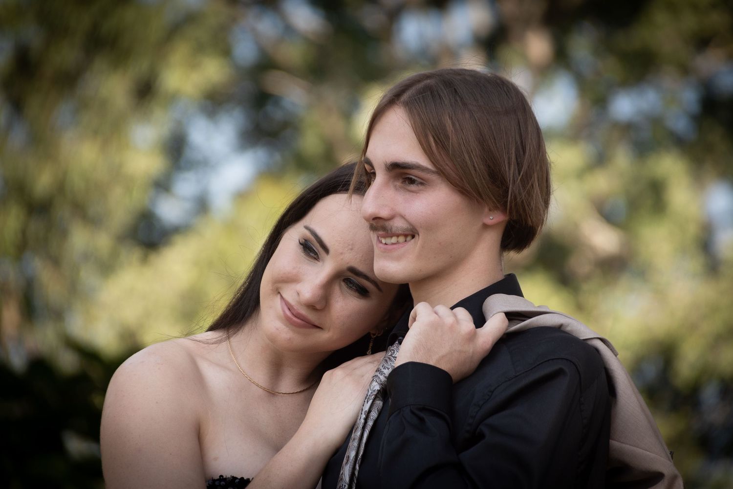 A loving couple share an intimate moment outdoors with soft natural lighting and blurred foliage in the background.