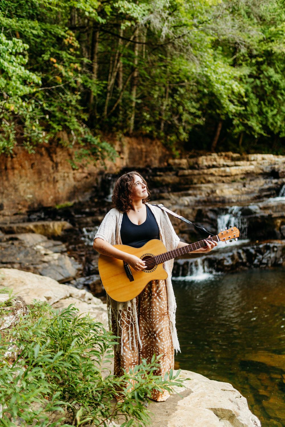 Curly haired woman playing acoustic guitar near a waterfall surrounded by lush greenery.