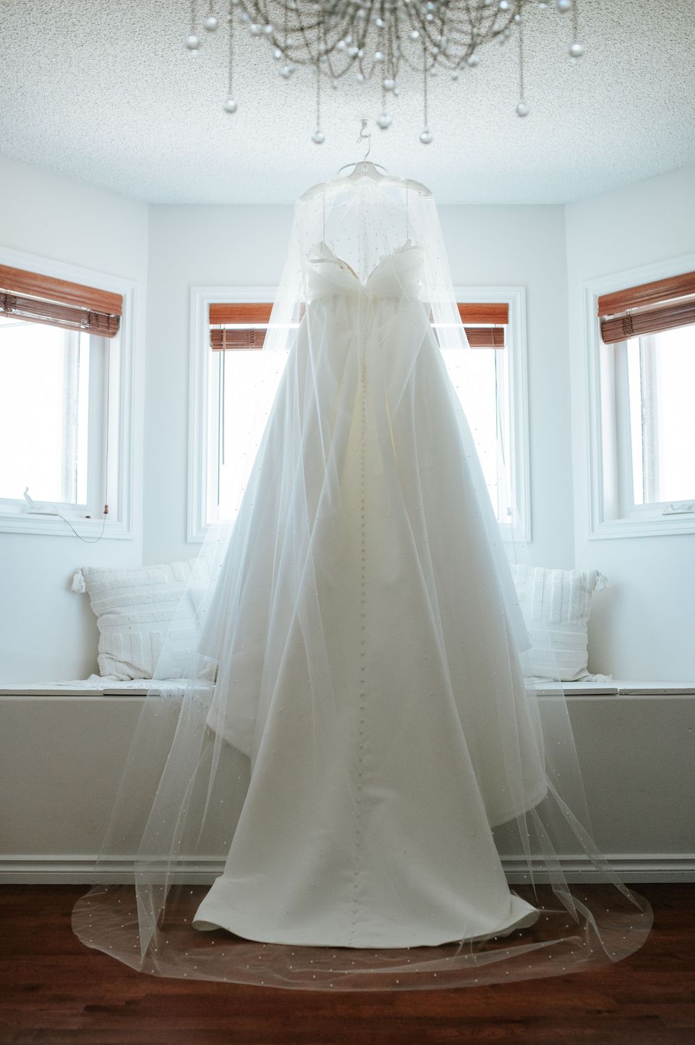 An elegant white wedding dress hangs from a crystal chandelier in a bright room with wooden blinds.