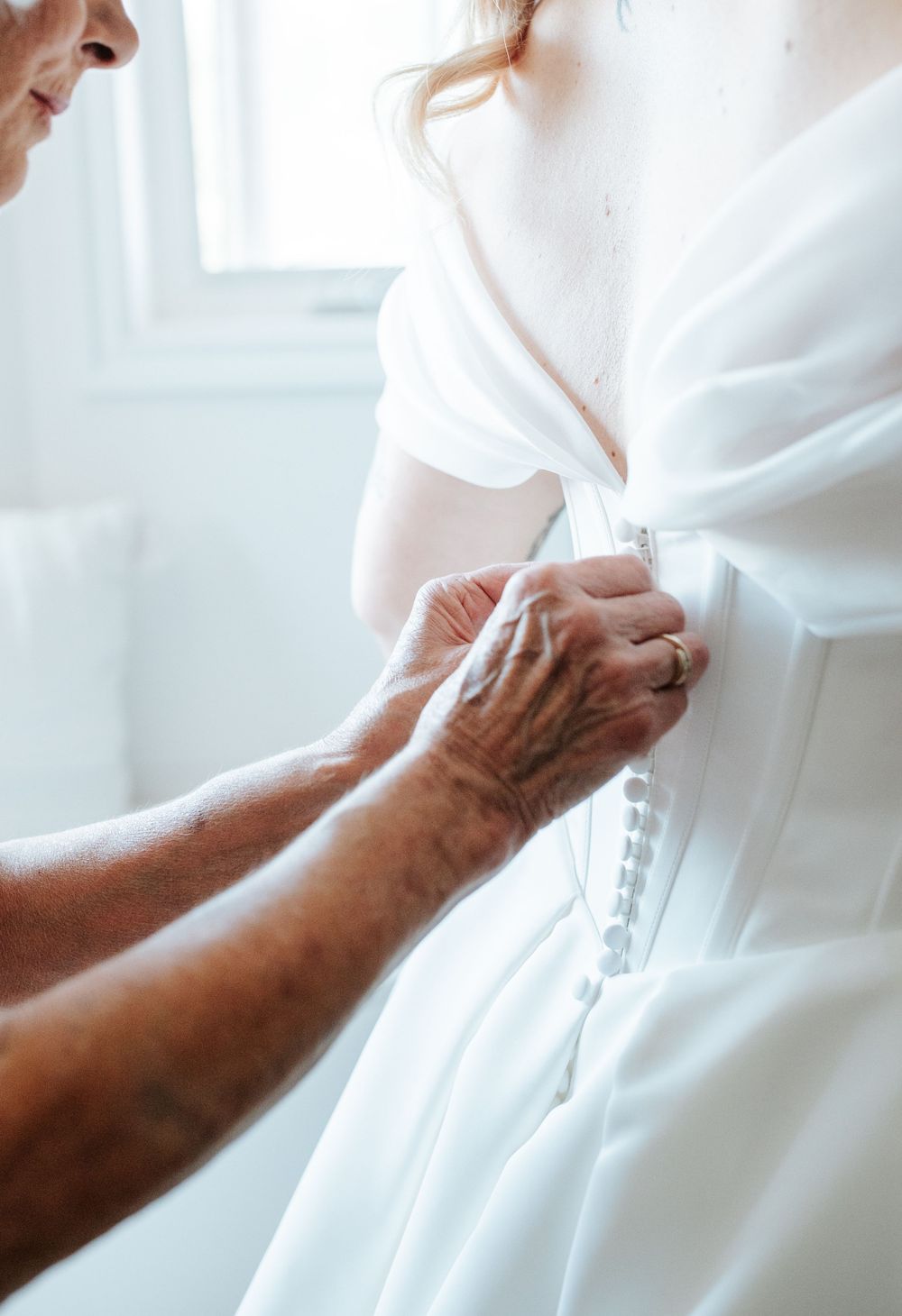 Close-up shot of hands fastening the back of a white wedding dress.