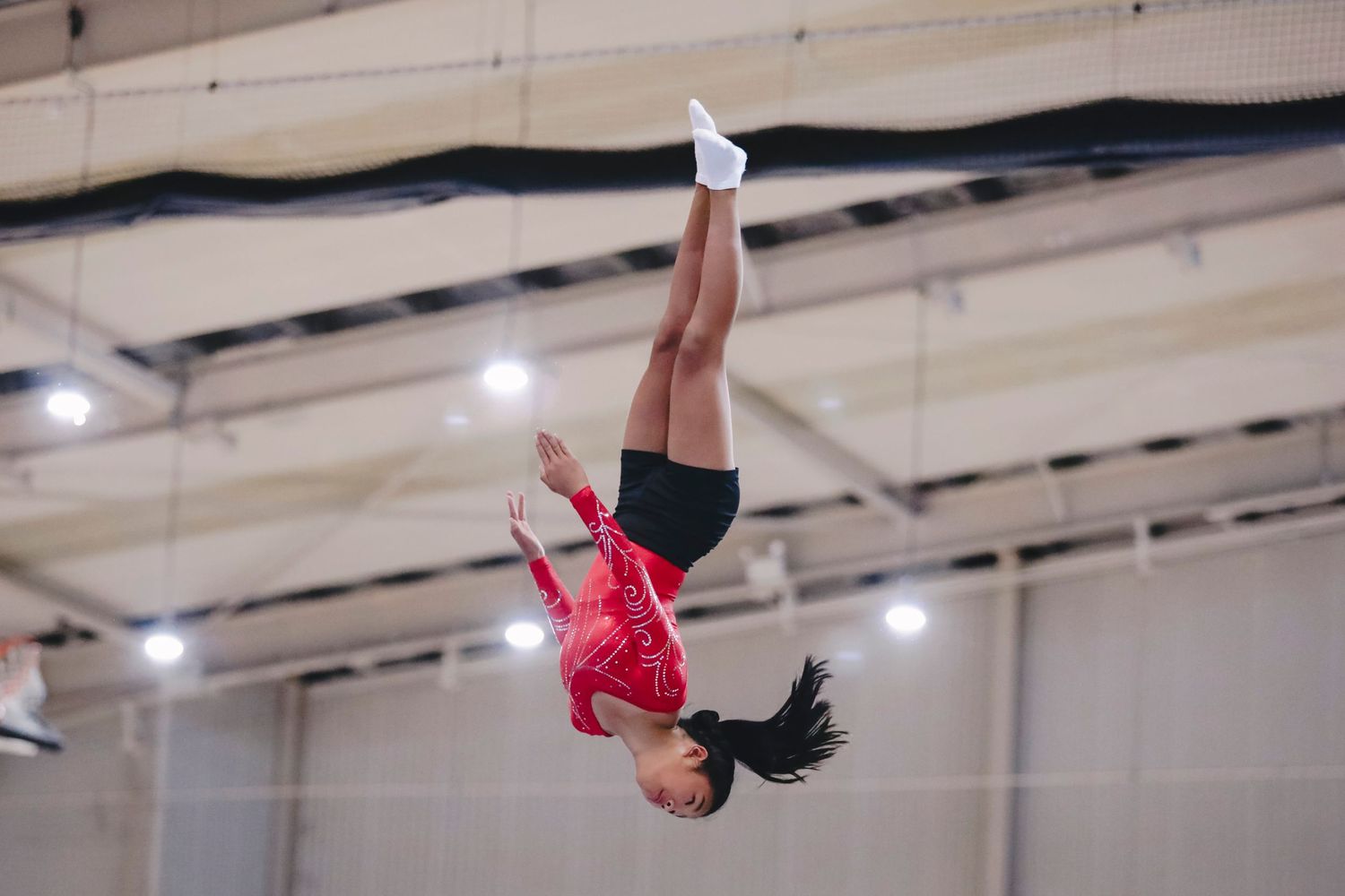 Gymnast in red leotard executing an inverted aerial maneuver during competition.