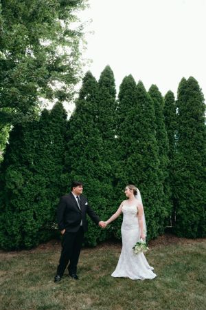 Bride and groom stand apart holding hands and looking at each other in front of tall evergreen trees at kansas city wedding