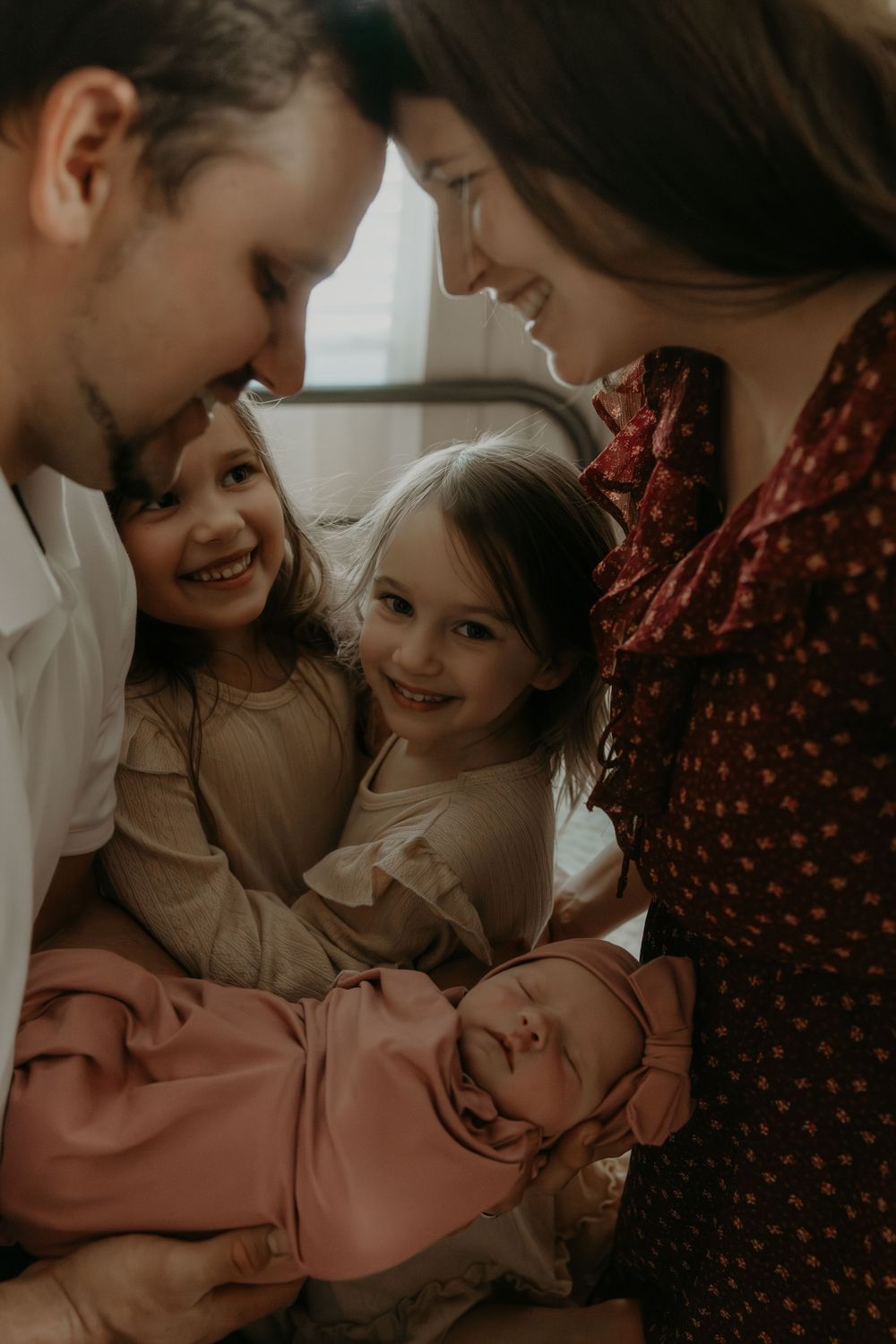 Family of four admiring their newborn baby wrapped in pink, two young girls smiling with parents.