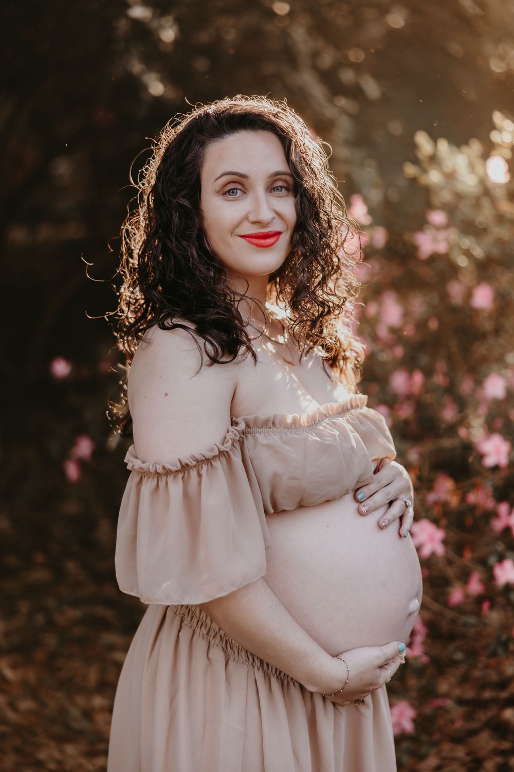 Pregnant woman in beige off-shoulder dress holding baby bump outdoors among pink flowers in golden sunlight.