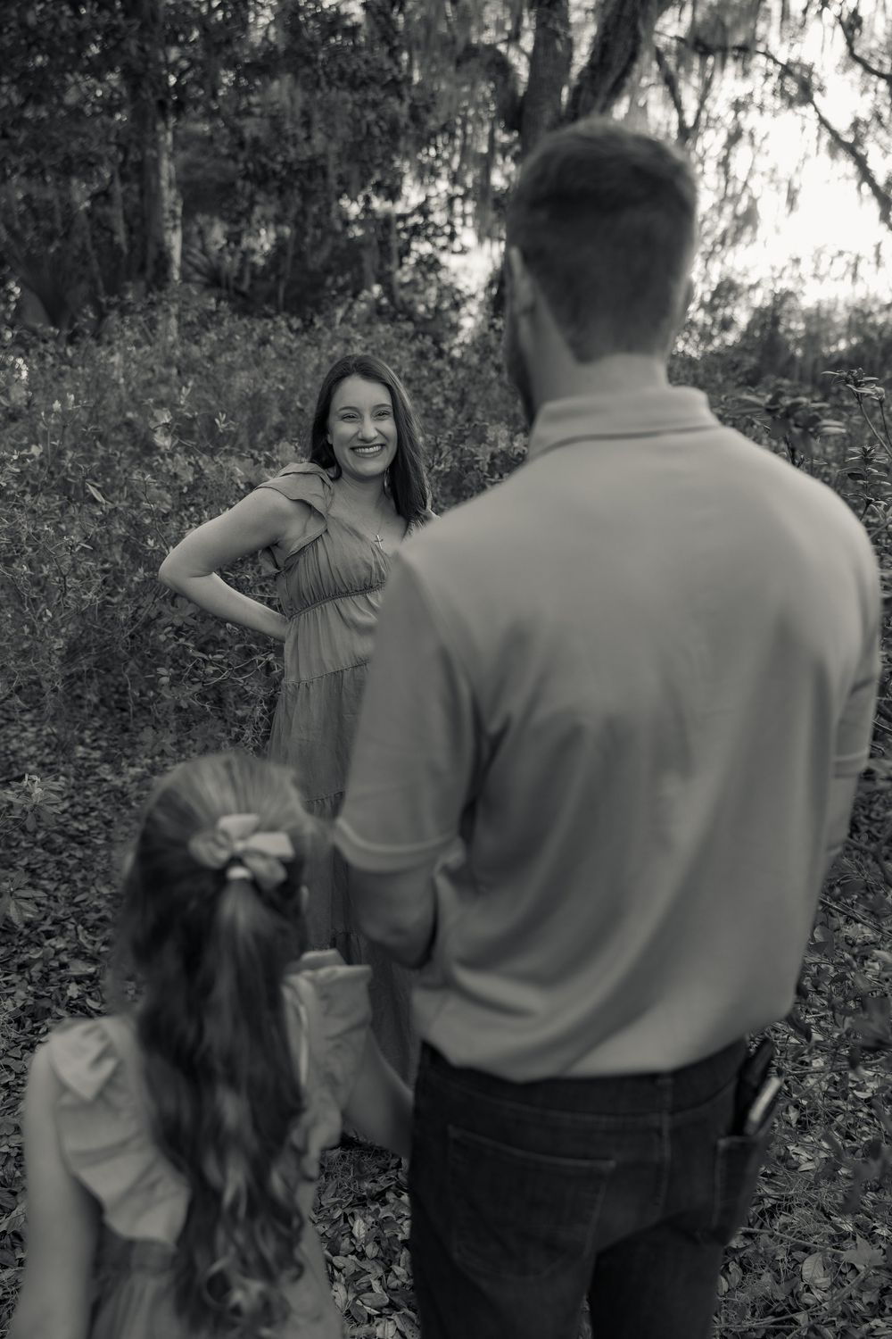 Black and white family photo of smiling pregnant woman with husband and daughter in wooded outdoor setting.