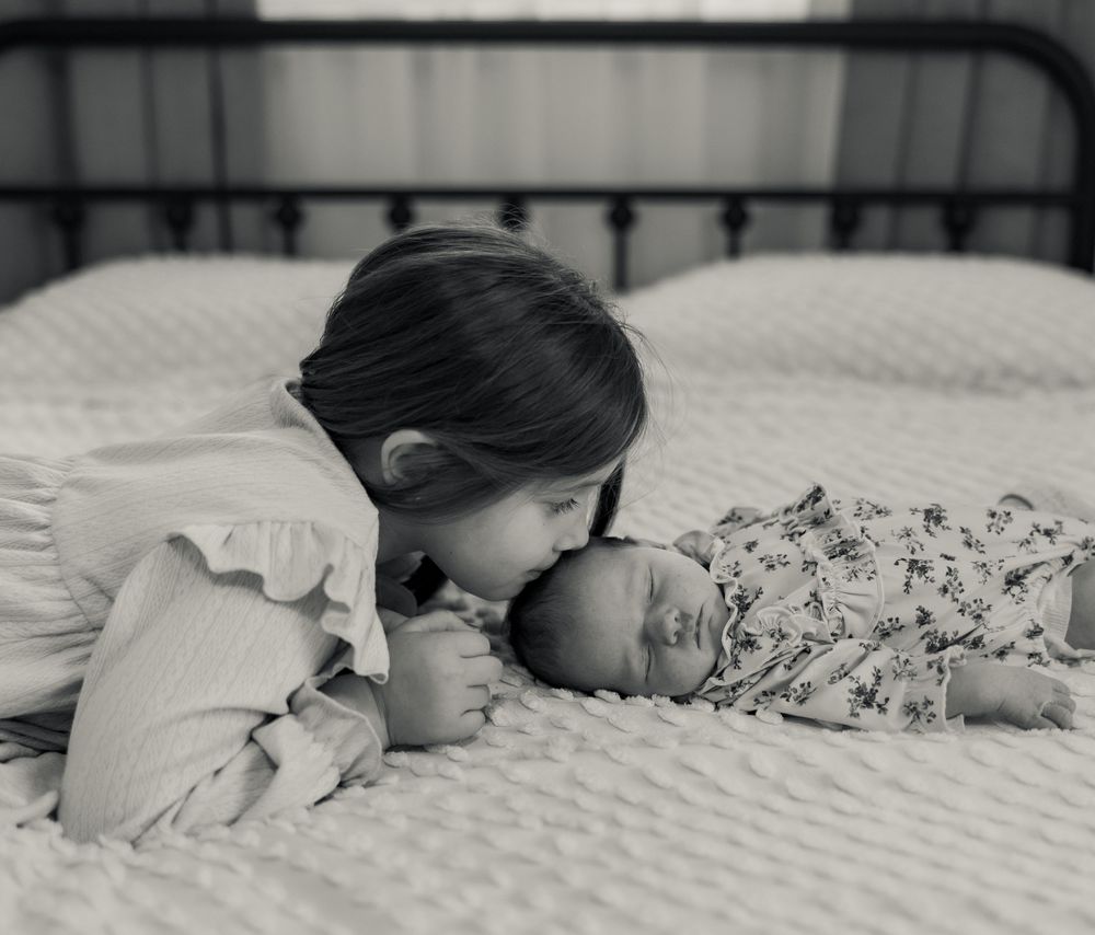 Black and white photo of a young girl gently kissing a newborn baby sibling on a bed.
